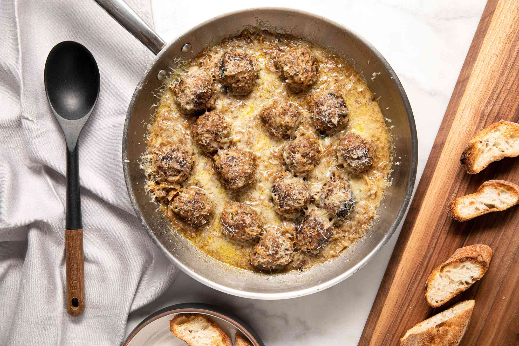 French Onion Meatballs in a Pan at a Table Setting With Toasted Baguette Slices on a Wooden Platter and a Spoon on the Counter, All on a White Table Cloth