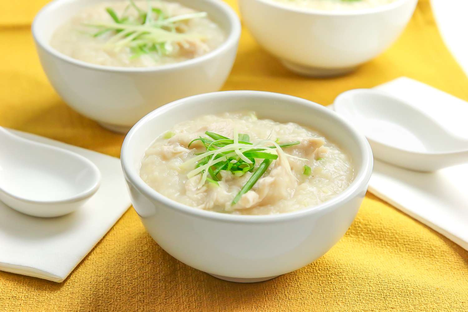 Bowl of Chicken Congee Topped with Sliced Ginger and Green Onions, Next to Soup Spoons and More Bowls of Congee, All on a Mustard Colored Kitchen Towel