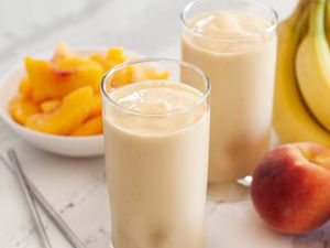 Two peach almond smoothies with whole bananas, peaches, and a plate of sliced peaches set around the smoothies.