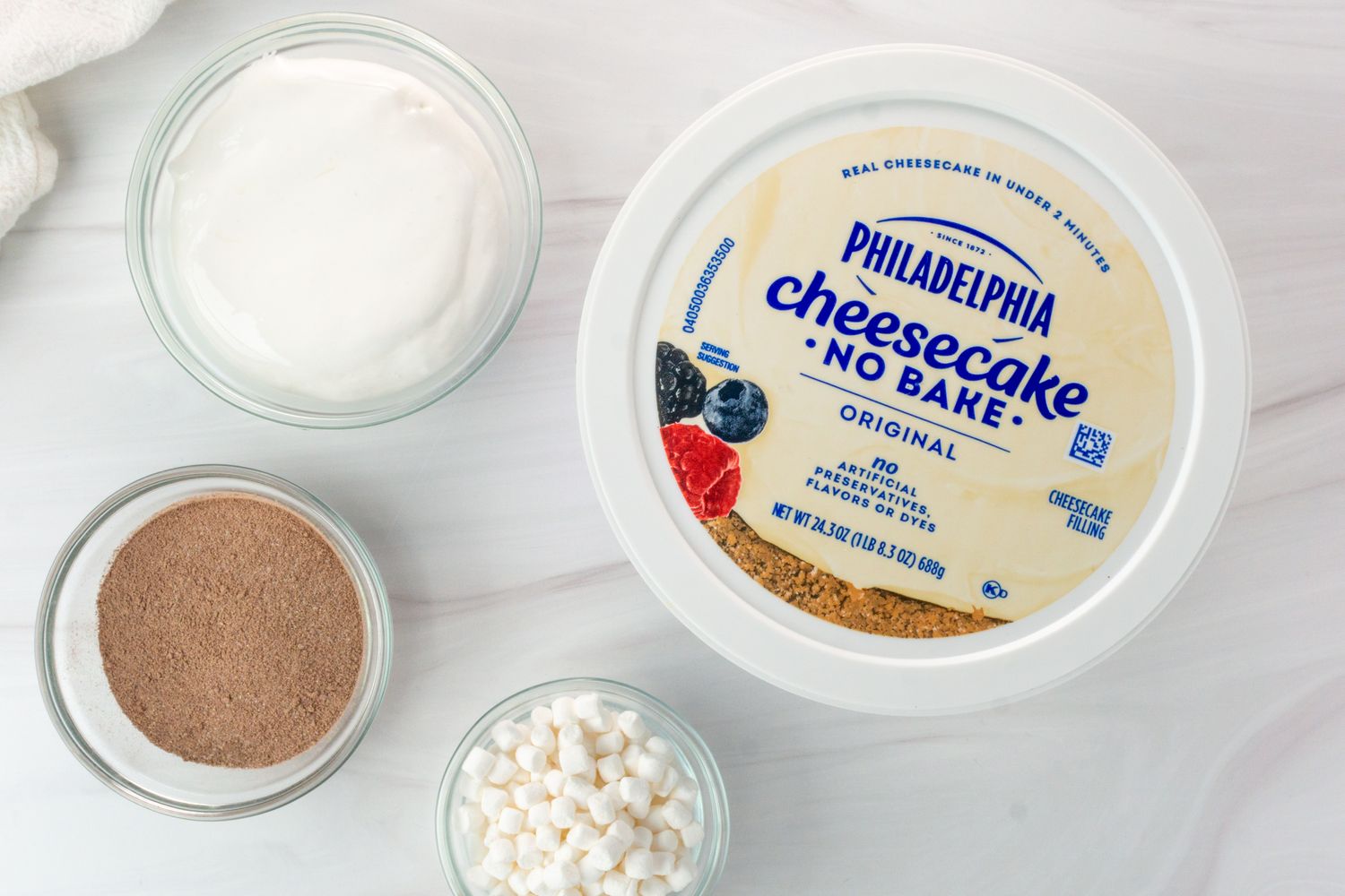 Ingredients for a hot cocoa dip including a tub of Philadelphia Cheesecake No Bake small bowls of cocoa powder mini marshmallows and whipped topping