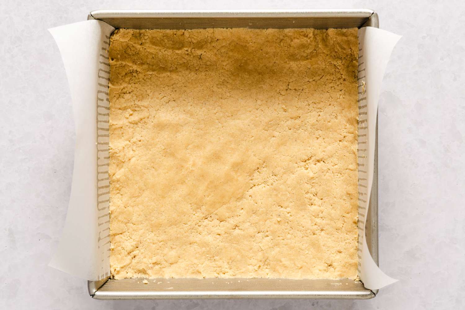 A baking dish lined with parchment paper and filled with a pressed crust mixture for caramel apple bars recipe before baking