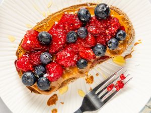 Slice of toast topped with peanut butter, raspberries, and blueberries, placed on a white plate with a fork to the side