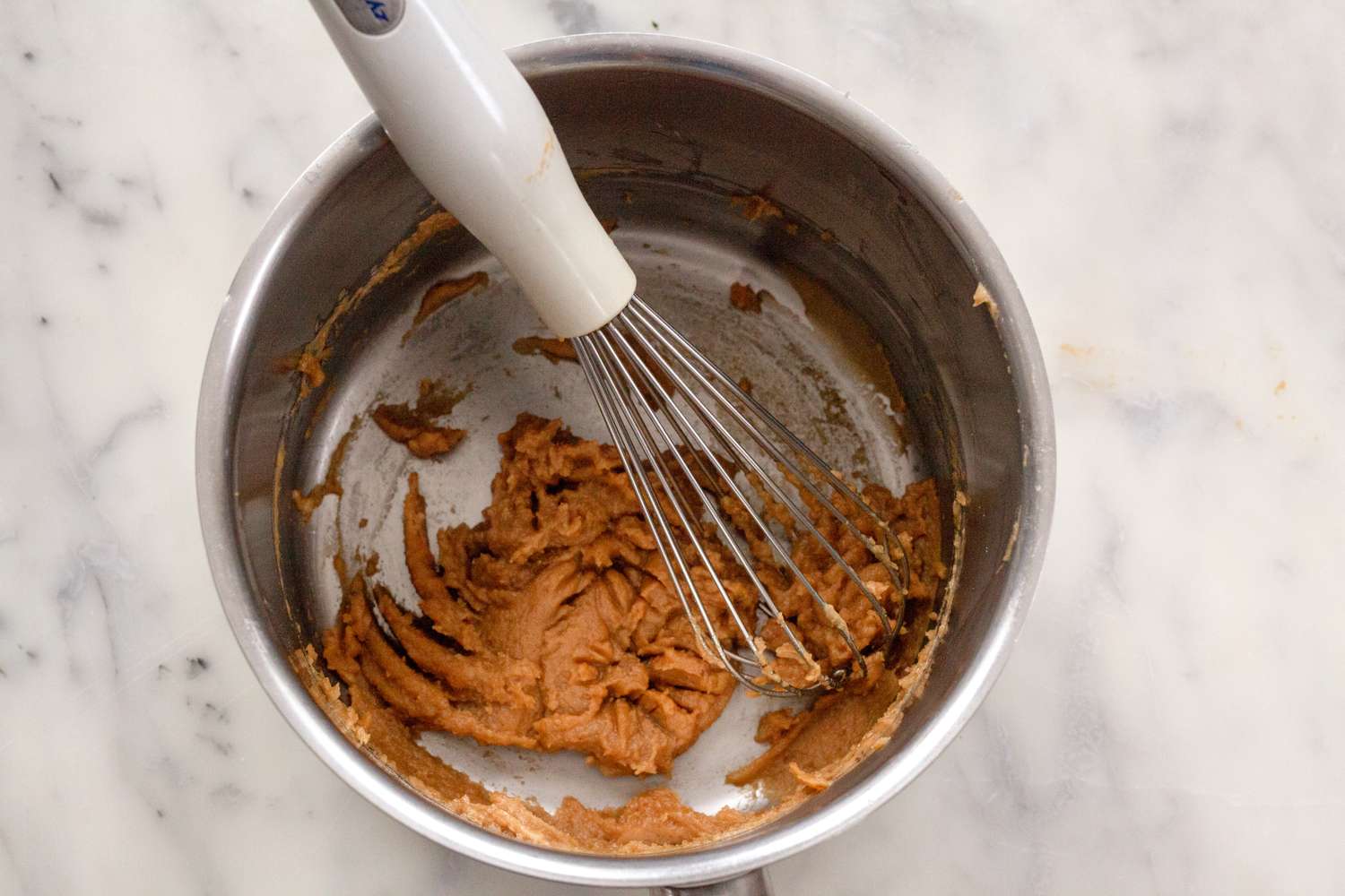 A saucepan with a whisk stirring a roux mixture on a marble surface
