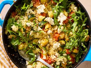 A skillet containing couscous, arugula, roasted vegetables, and cheese crumbles, with a serving spoon
