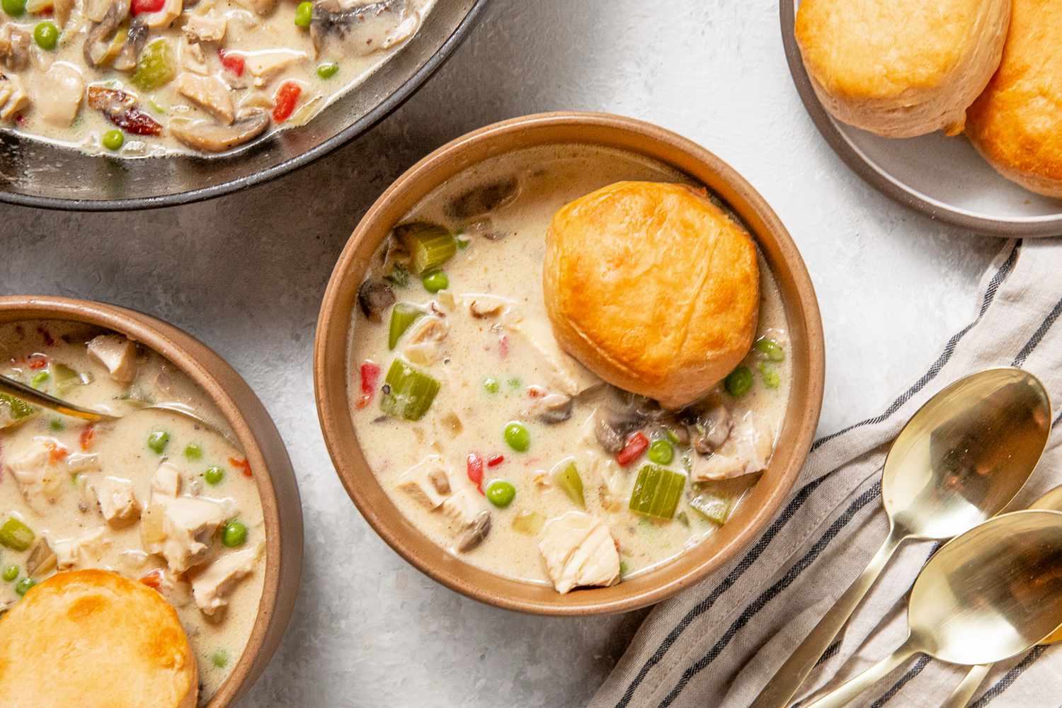 Chicken a la king in a bowl with a biscuit at a table setting with another serving in a bowl with a biscuit, a bowl of biscuits, and a pot with more chicken a la king
