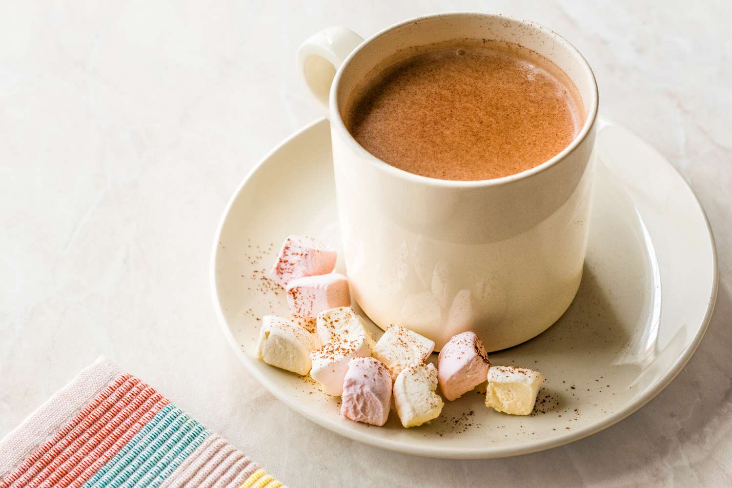 A cup of hot cocoa on a saucer with marshmallows placed next to a striped fabric