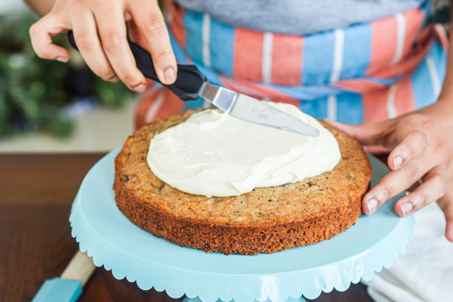 A person in a striped apron spreading frosting on a layer of hummingbird cake on a cake stand.