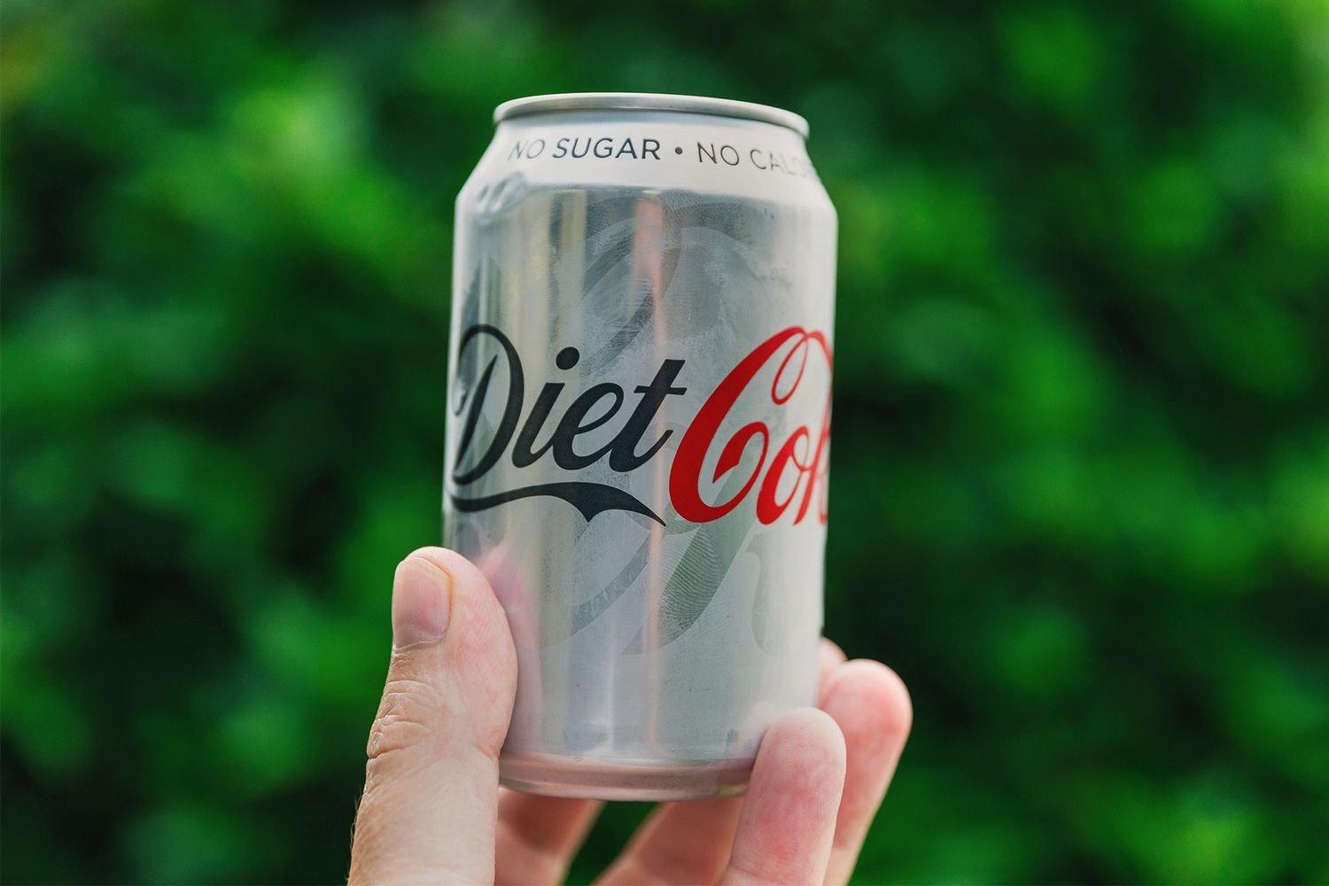 A person holding a can of Diet Coke with a blurred foliage background