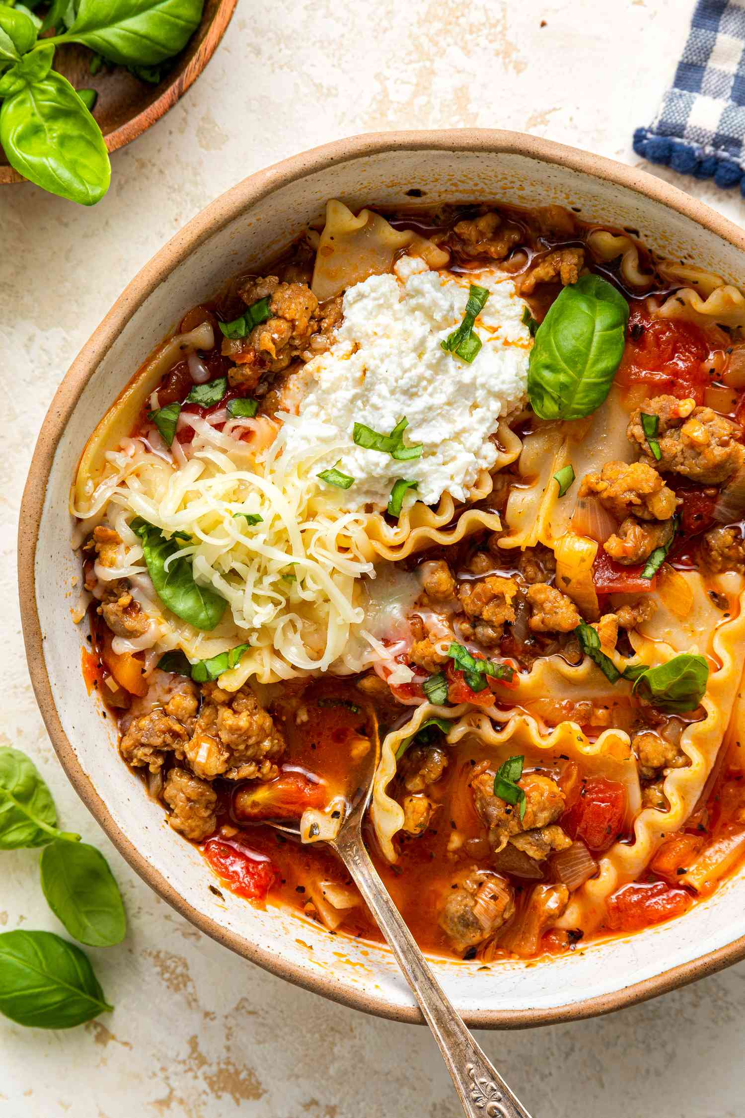 Lasagna Soup in a Bowl With Noodles, Sausage, Ricotta, and Fresh Basil, and in the Surroundings, a Saucer With Basil Leaves, Basil Leaves Free Flowing on the Counter, and a Blue and White Checkered Kitchen Towel 