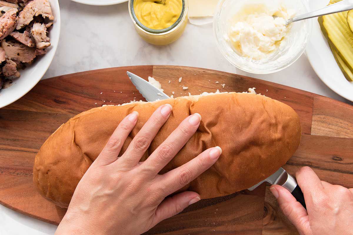 A loaf of french bread being sliced horizonally with a chefs knife. One hand rests on top of the loaf. Shredded pork, a glass container of mustard, a small glass bowl of mayonnaise and a partial plate of sliced pickles is visible above.