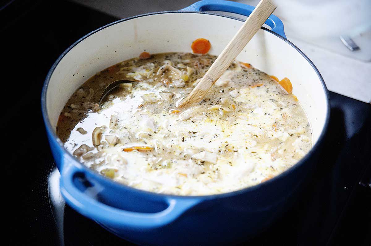A blue cast iron dutch oven with easy to make homemade creamy chicken noodle soup and cream stirred into it.