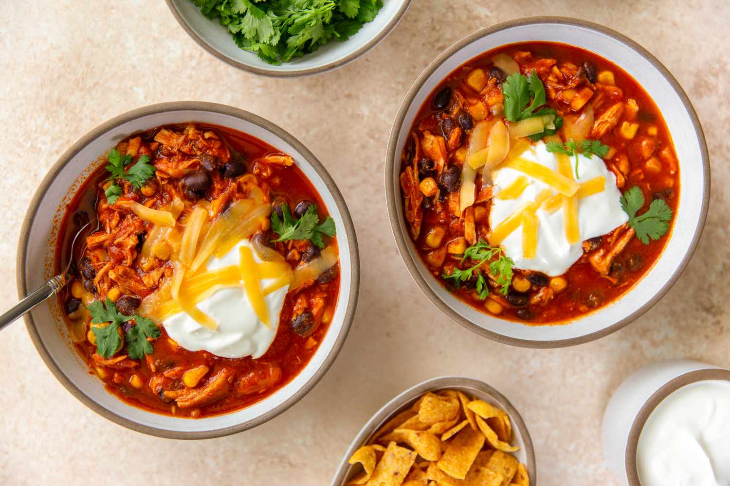 Bowls of chicken chili topped with sour cream and shredded cheese garnished with parsley and served with chips and condiments