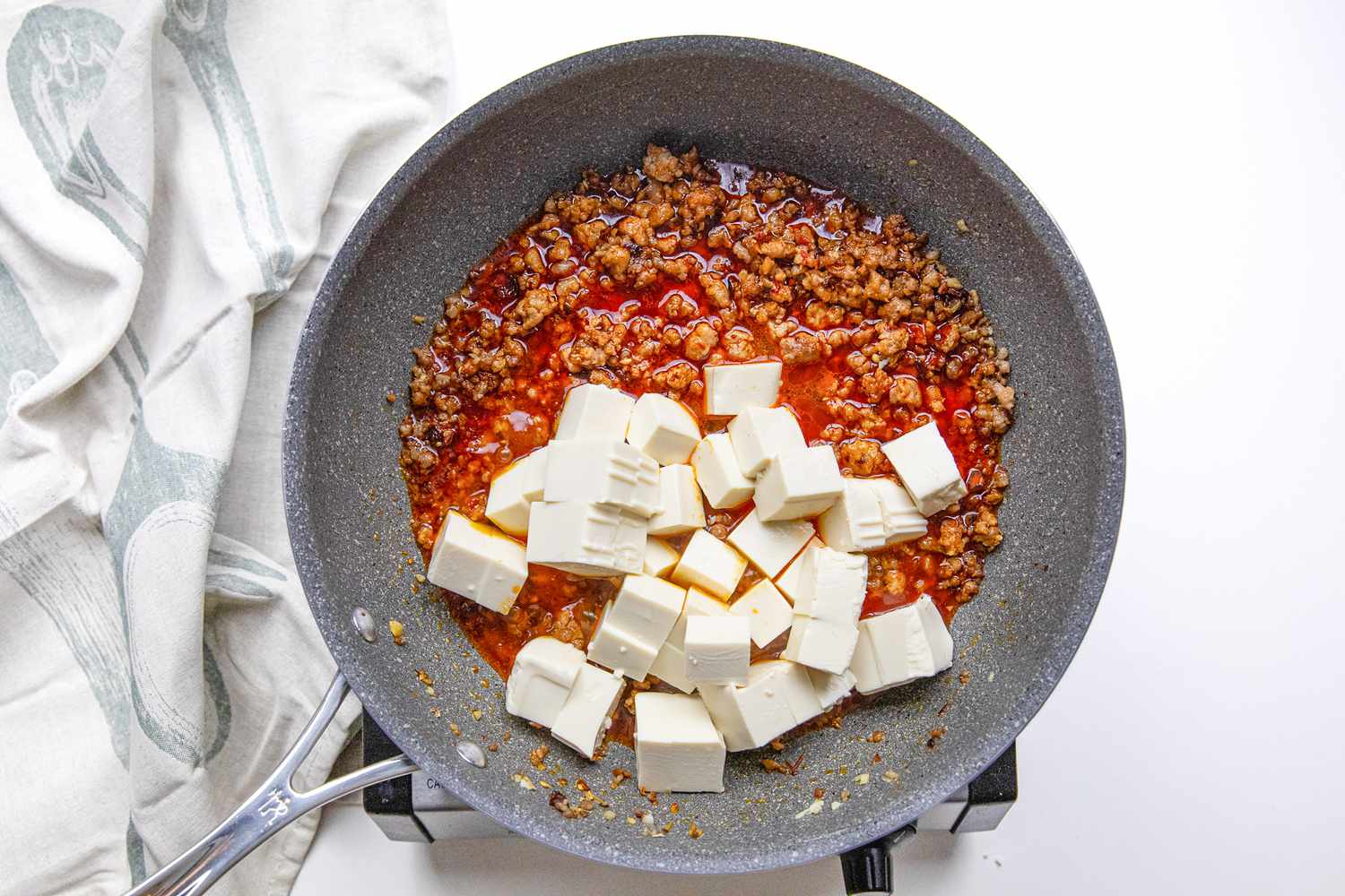 Tofu Cubes and Broth Added to Pan on a Portable Burner Next to a White Kitchen Towel for Sichuan Tofu Recipe