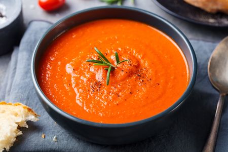 A bowl of tomato soup garnished with fresh rosemary placed on a gray cloth with a piece of bread on the side