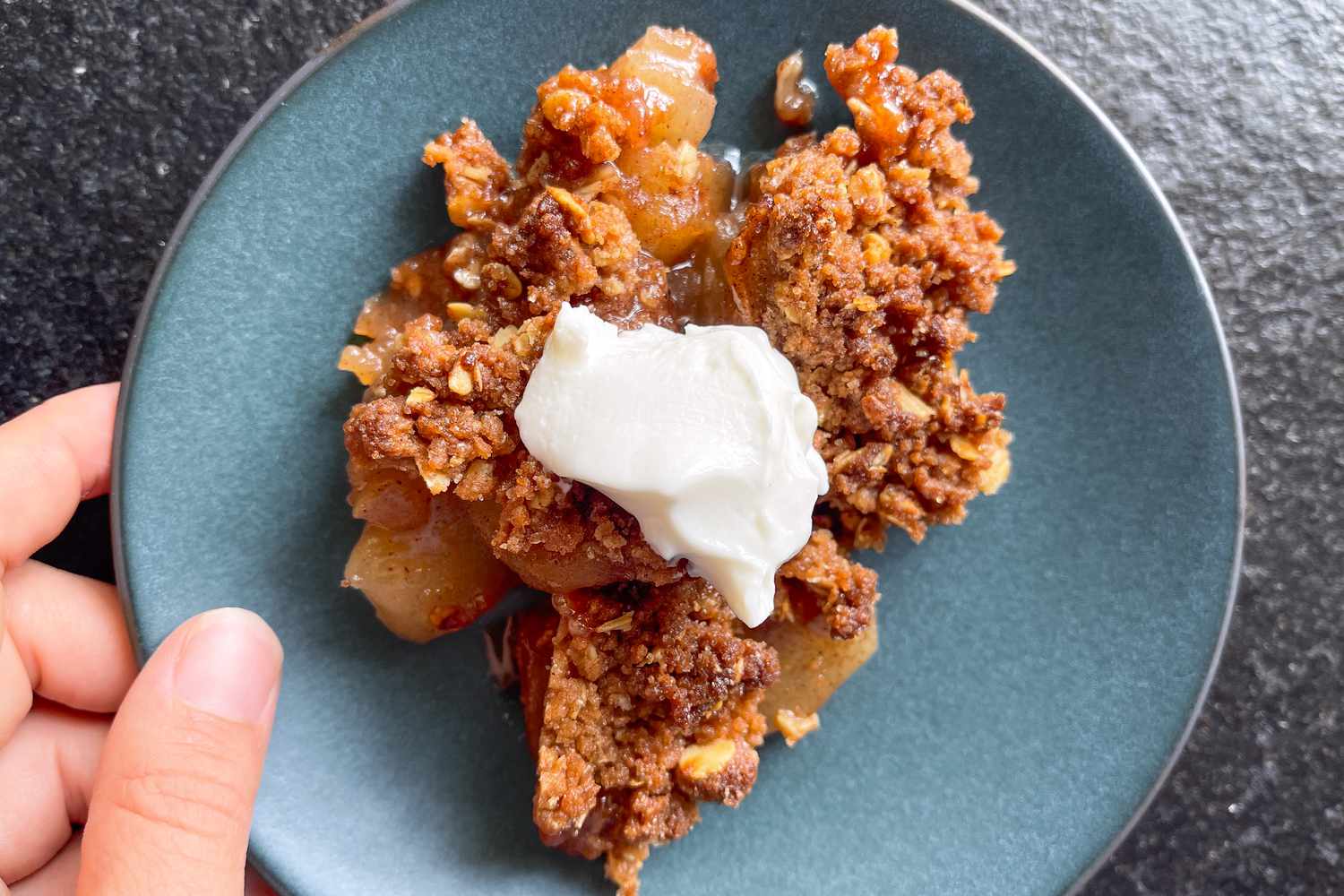 A hand holding a small plate with a serving of apple crisp on it, topped with a little whipped cream