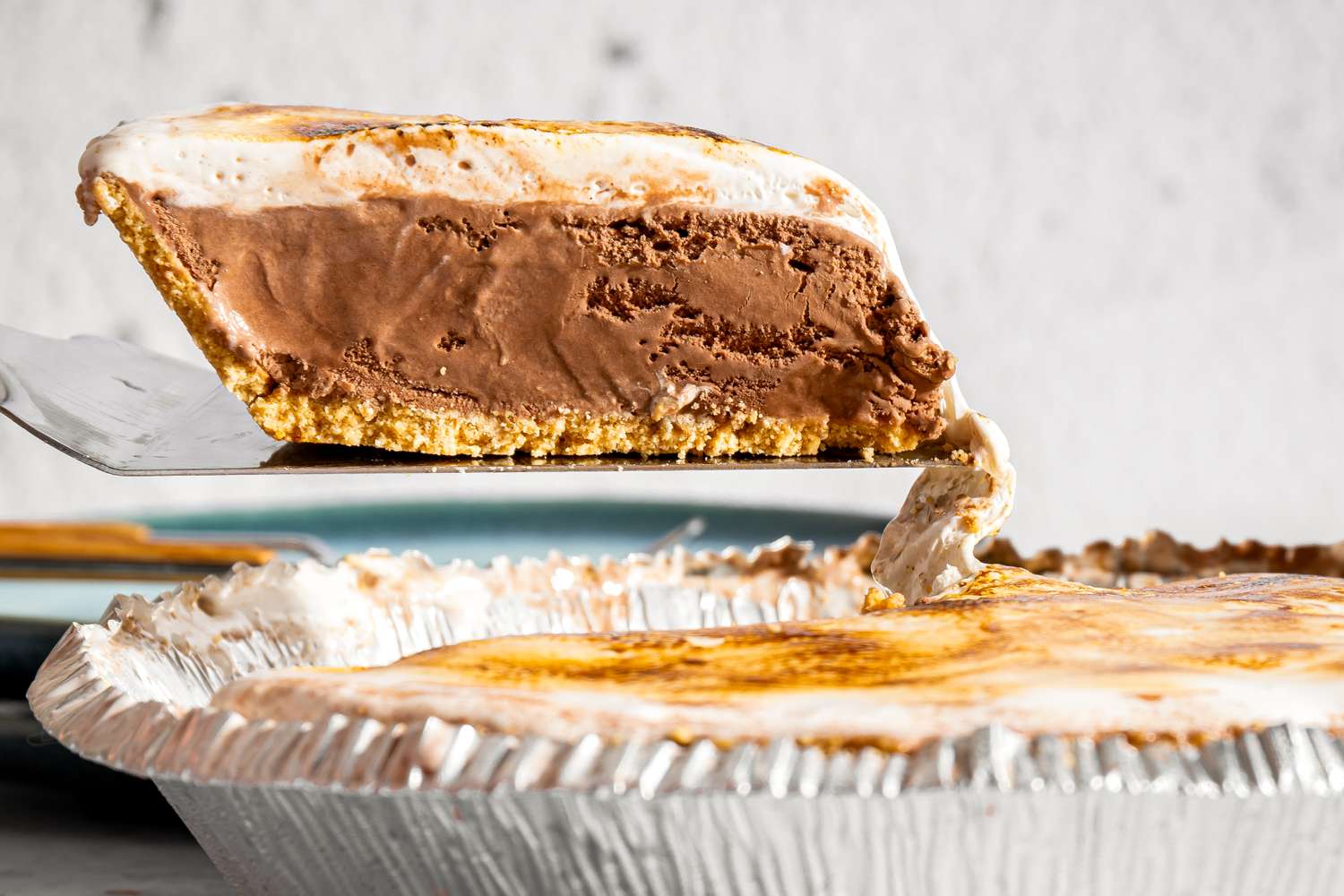 A slice of frozen smores pie being lifted from the tin showing layers of graham cracker crust chocolate and marshmallow topping