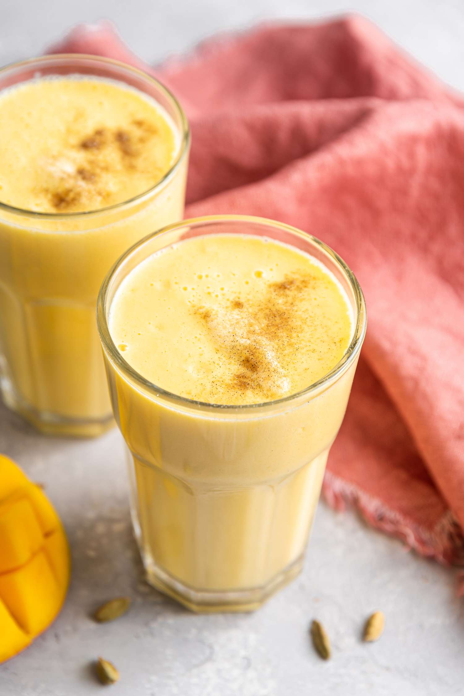 Easy Mango Lassi Surrounded by Another Glass, Kitchen Towel, and Cardamom Pods