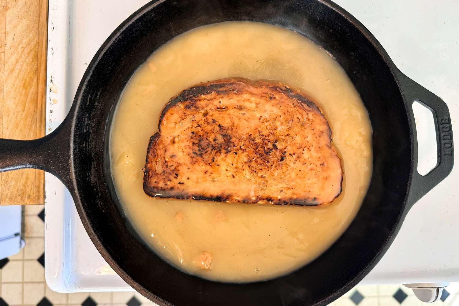 A piece of bread soaking in liquid within a cast iron skillet on a stovetop surface