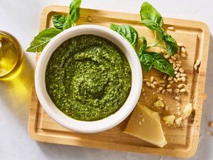 Bowl of pesto on a wooden cutting board with basil leaves pine nuts Parmesan cheese and olive oil nearby