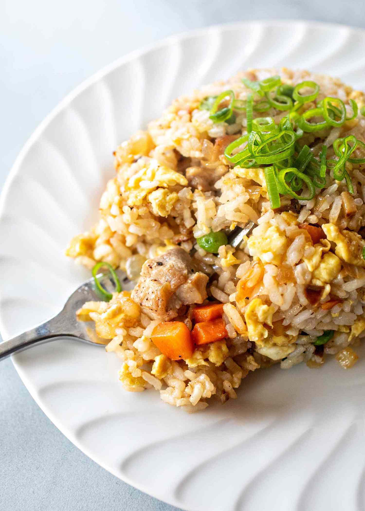 Scalloped plate with pork fried rice scooped on it. Chopped scallions, peas and carrots are visible in the rice. A fork is under the rice on the left side.
