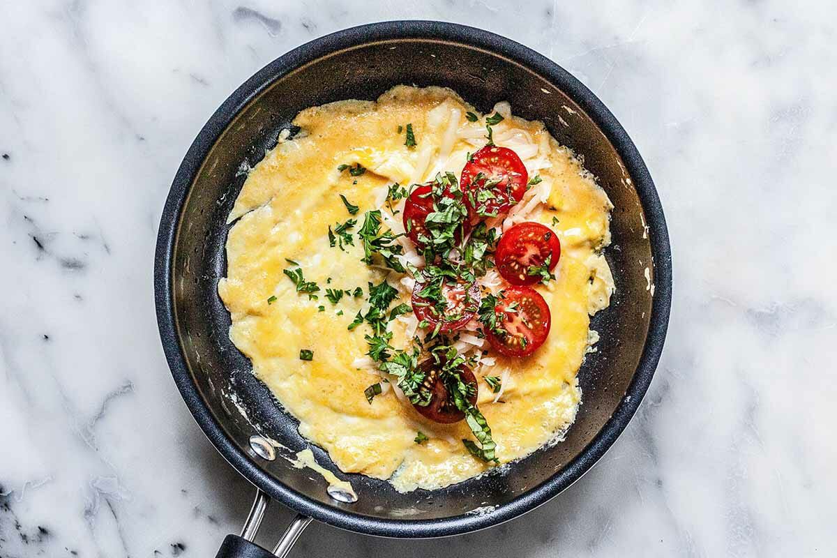 Overhead of an open omelette cooking in a non-stick pan with cherry tomatoes and chopped parsley in the center 