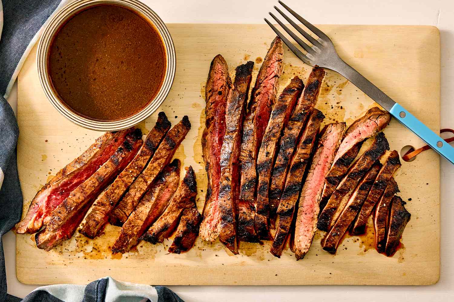 Sliced cooked flank steak on a wooden cutting board with a bowl of sauce and a fork