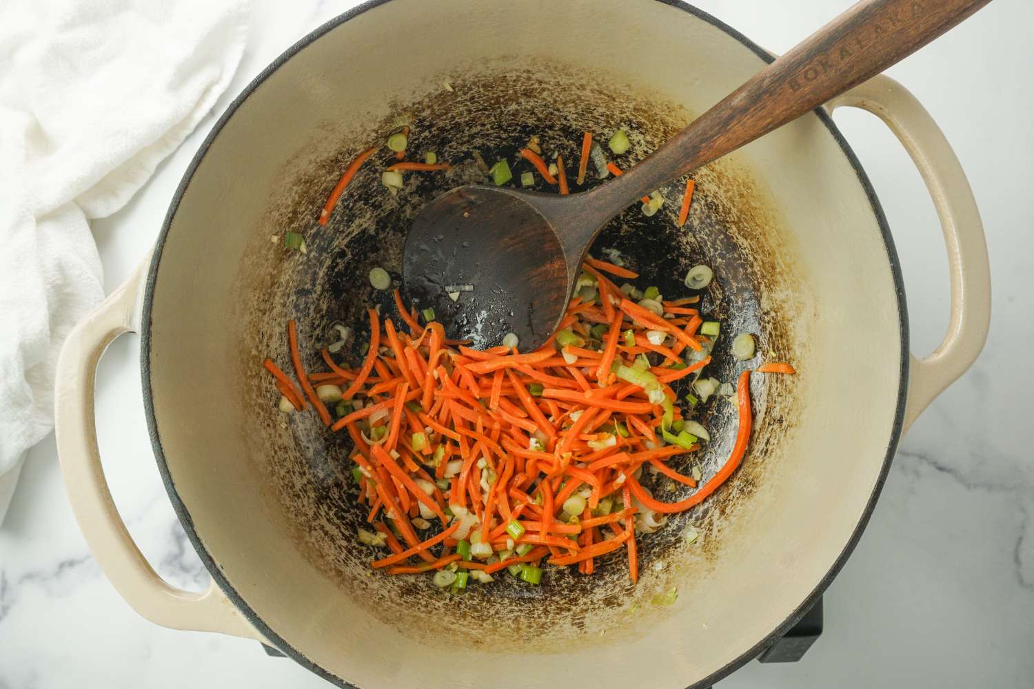 A pot with shredded carrots and sliced green onions being sauted a wooden spoon rests in the pot