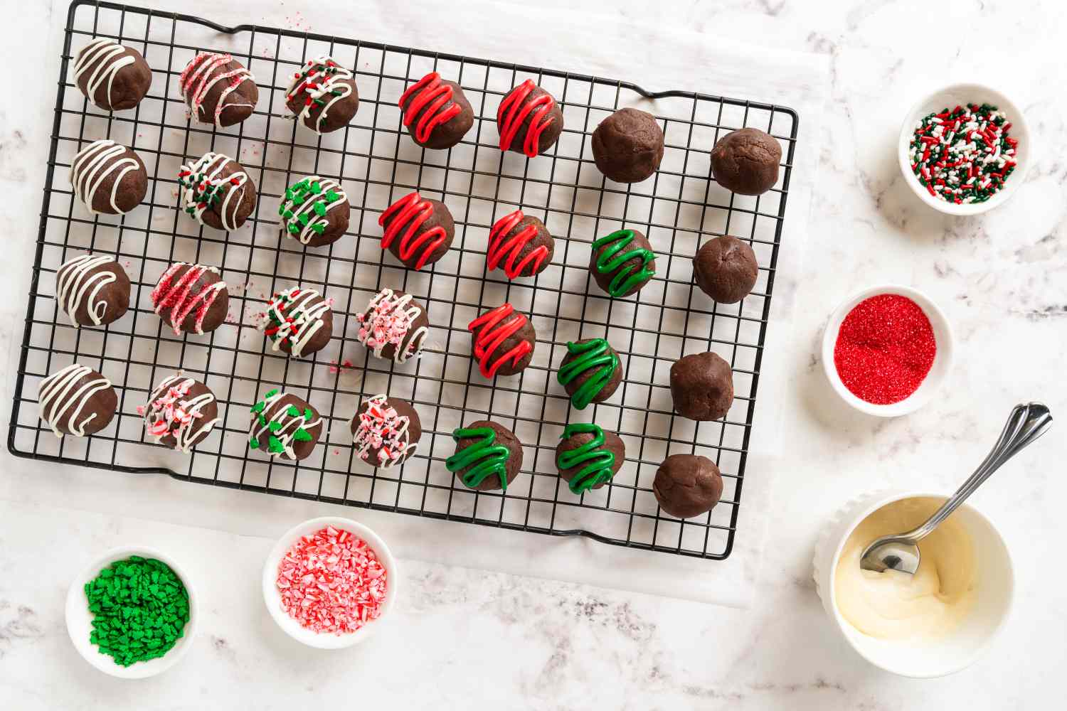 Decorative chocolate bonbons with colorful drizzles and sprinkles on a wire cooling rack surrounded by bowls of sprinkles and melted chocolate
