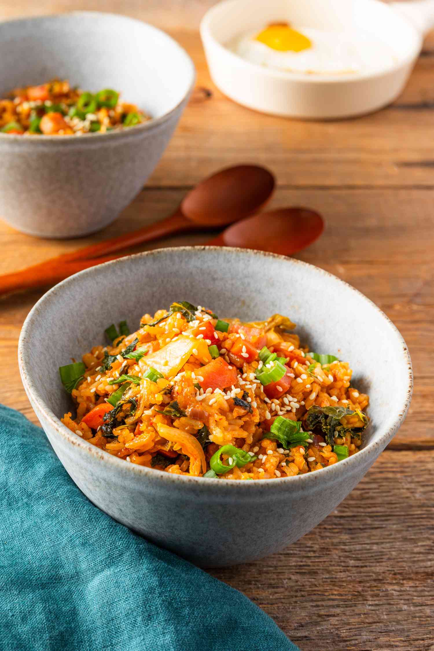 Bowl of kimchi and spam fried rice at a table setting with another seving in a bowl, a plate with a fried egg, two spoons on the counter, and a blue kitchen linen
