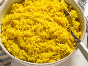 Closeup view of a large bowl of golden rice with a serving spoon all on marble countertop with a cloth napkin, forks, plates a glasses
