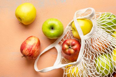 A white fabric mesh bag laying on a pink surface, with apples spilling out.