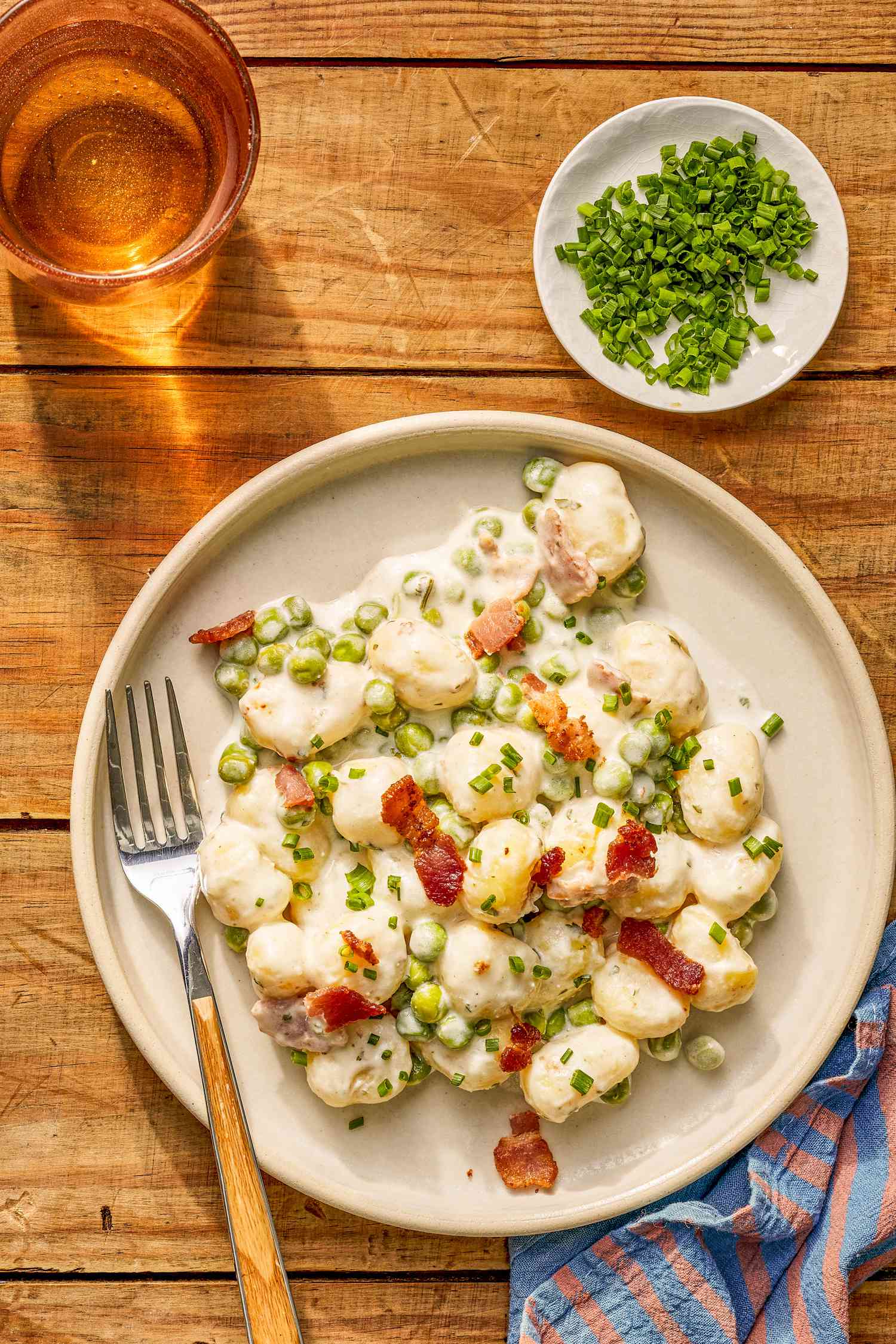 Plate of Creamy Skillet Gnocchi With Bacon and Peas at a table setting with a drinks, a small plate of chopped chives, and a blue and pink table napkin