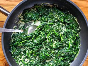 A skillet with Homemade Creamed Spinach and a serving spoon set on a wood table.