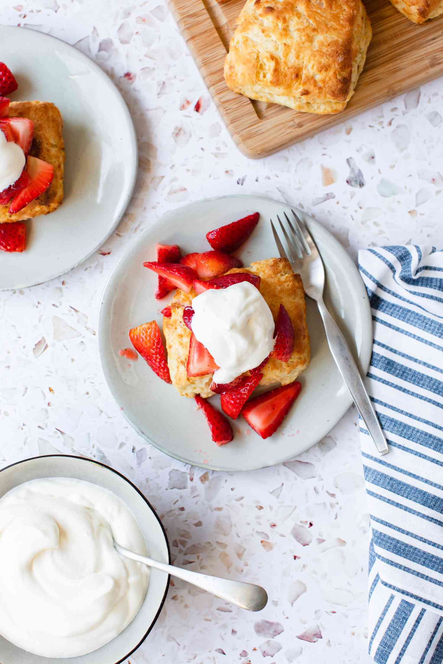Two Plates of Easy Strawberry Shortcake with a Fork Next to a Bowl of Whipped Cream and a Board of Scones