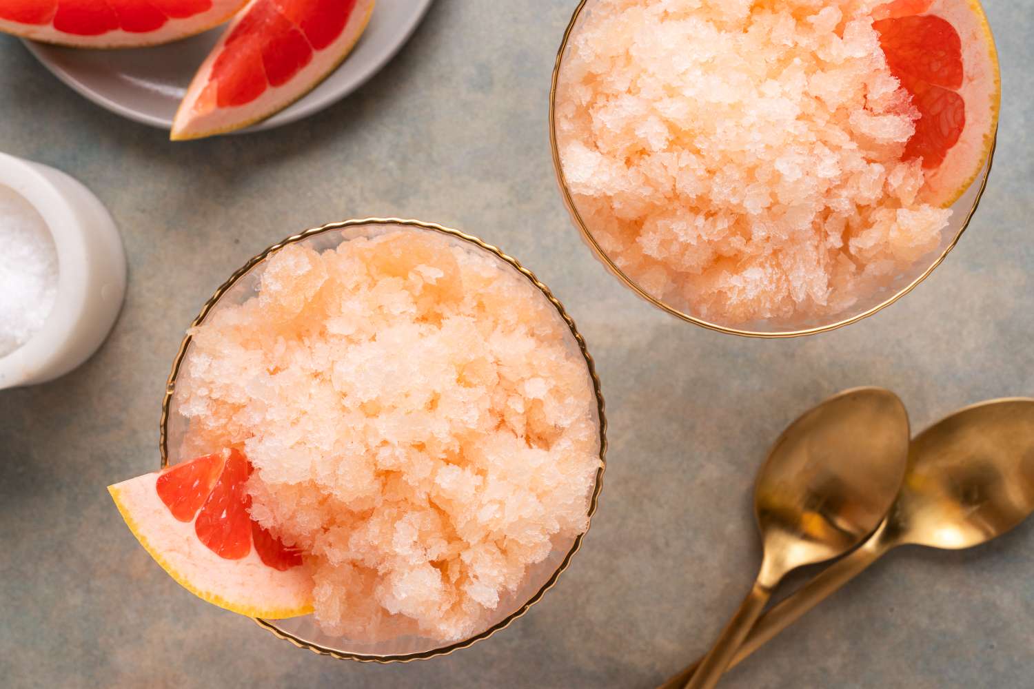 Two glasses of grapefruit granita garnished with grapefruit slices accompanied by gold spoons and a grapefruit plate in the background
