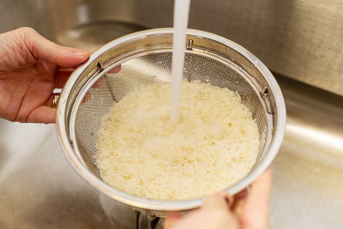 A strainer full of white rice under running water from a kitchen sink