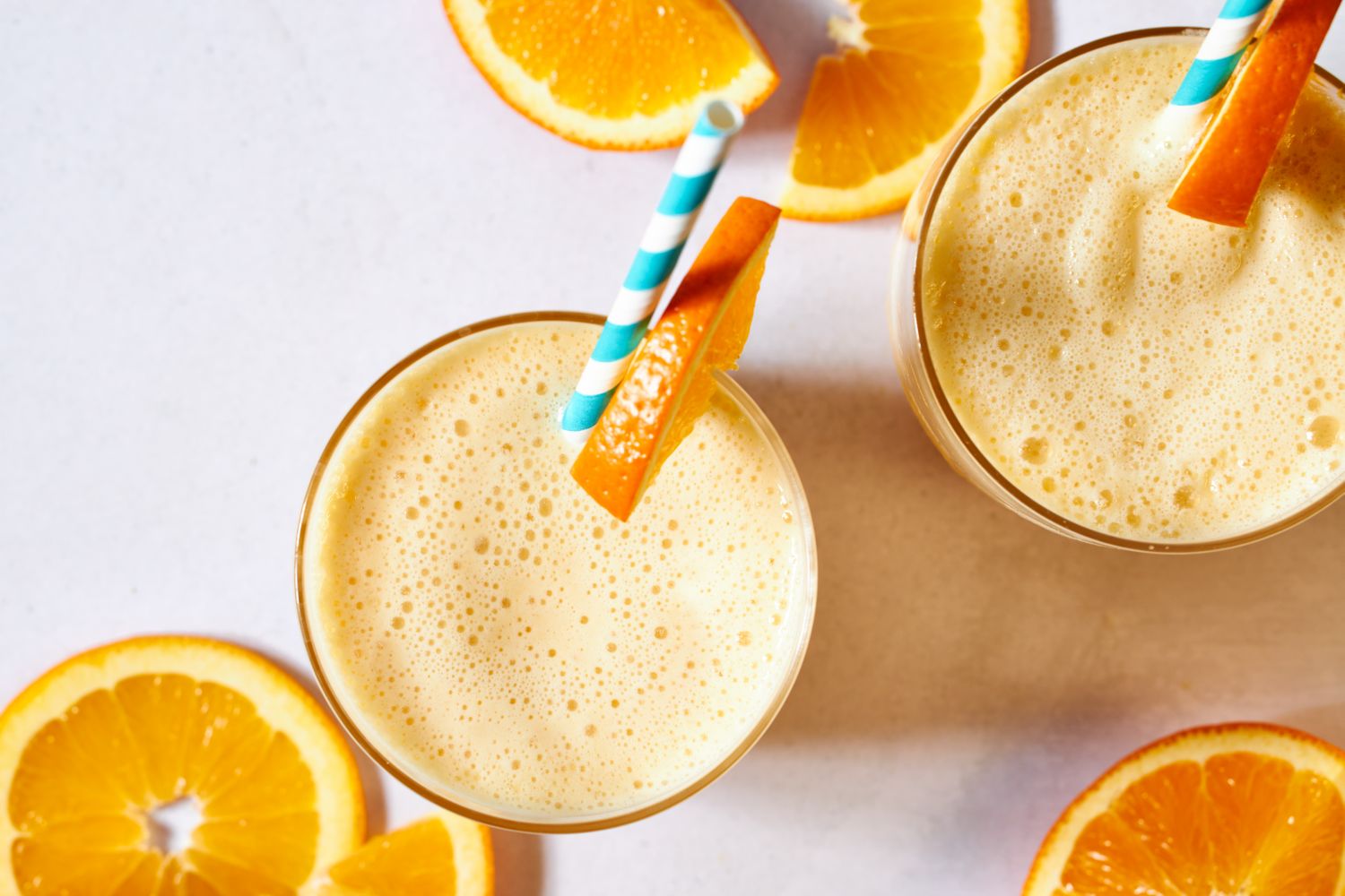 Two frothy orange beverages with striped straws, garnished with orange slices, surrounded by more orange slices on a light background