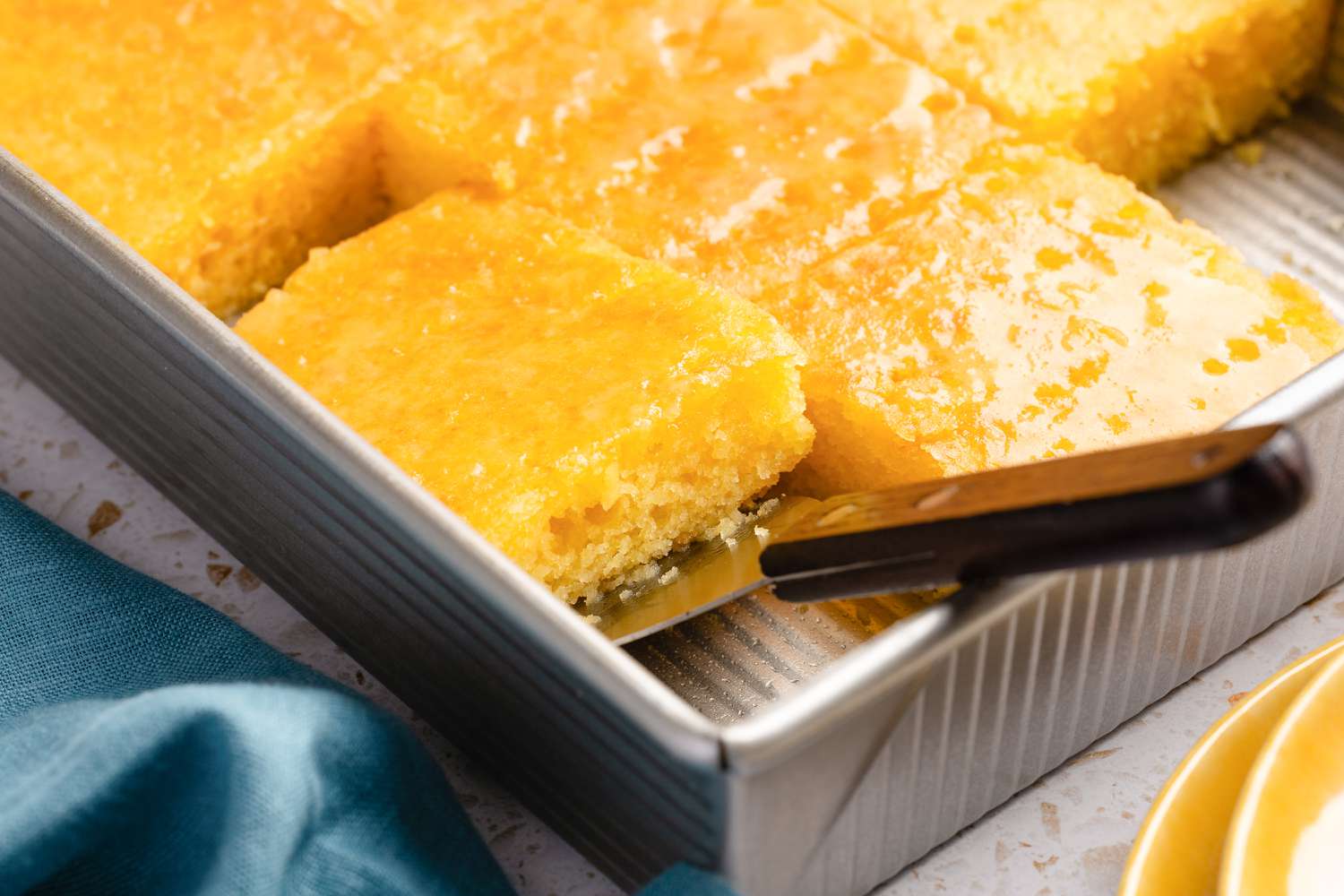 Angled view of a slice of orange poke cake being picked up by a spatula from a metal cake pan
