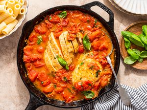 chicken pomodoro in a cast iron skillet at a table setting with a bowl of rigatoni, bowl of basil, stack of plates, and a table napkin