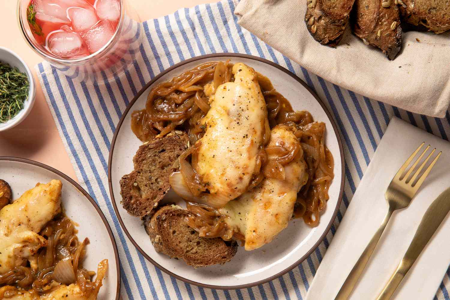 french onion chicken on a plate with a slice of bread at a table setting with another serving on a plate, a glass, a small bowl of herbs, and a bowl of sliced bread
