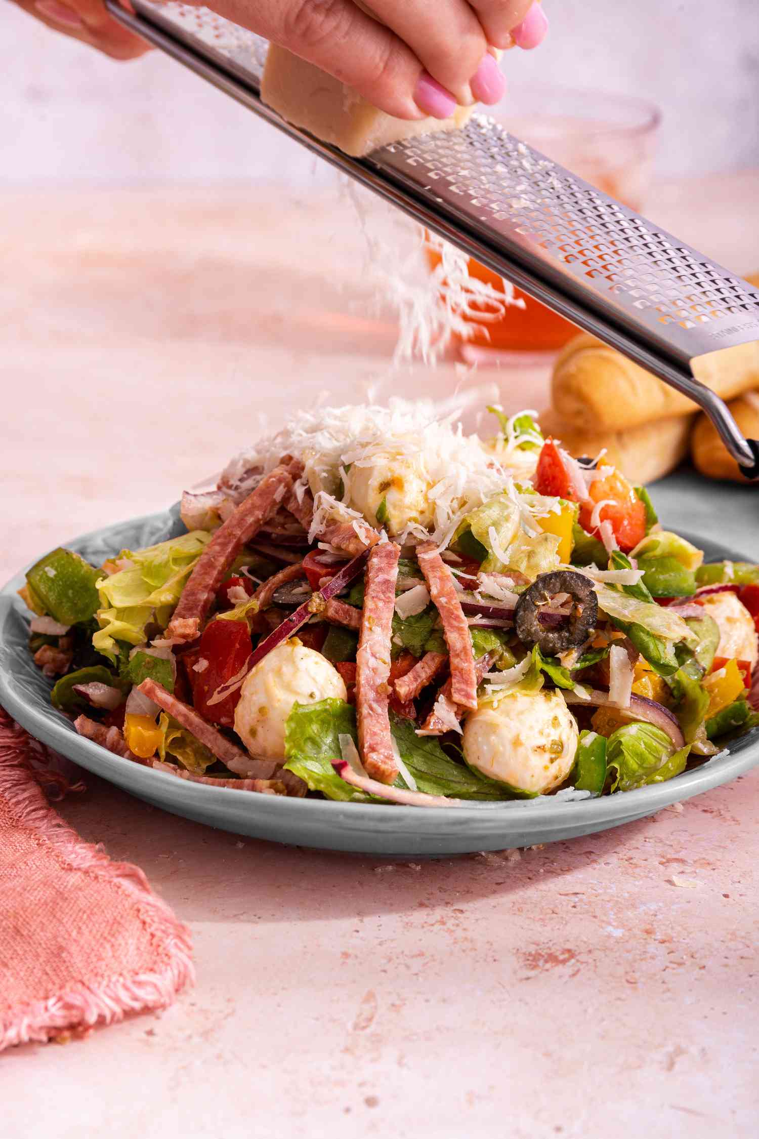 Parmesan Grated Onto a Serving of Pizza Salad on a Plate Next to a Plate of Bread Sticks, a Drink, and a Pink Kitchen Towel