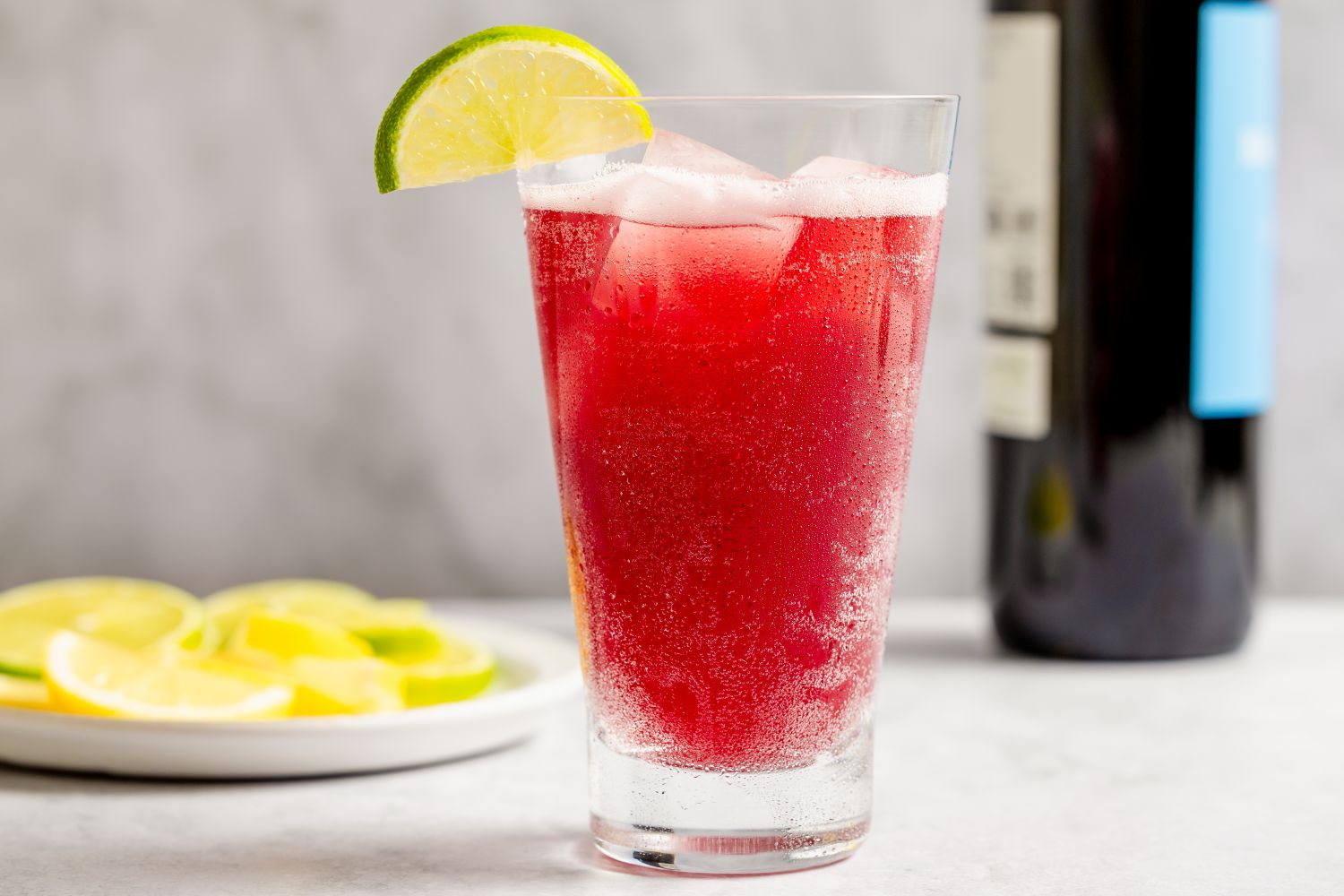 Tall glass of a red spritz cocktail garnished with a lime wedge plate of lime slices and a wine bottle in the background
