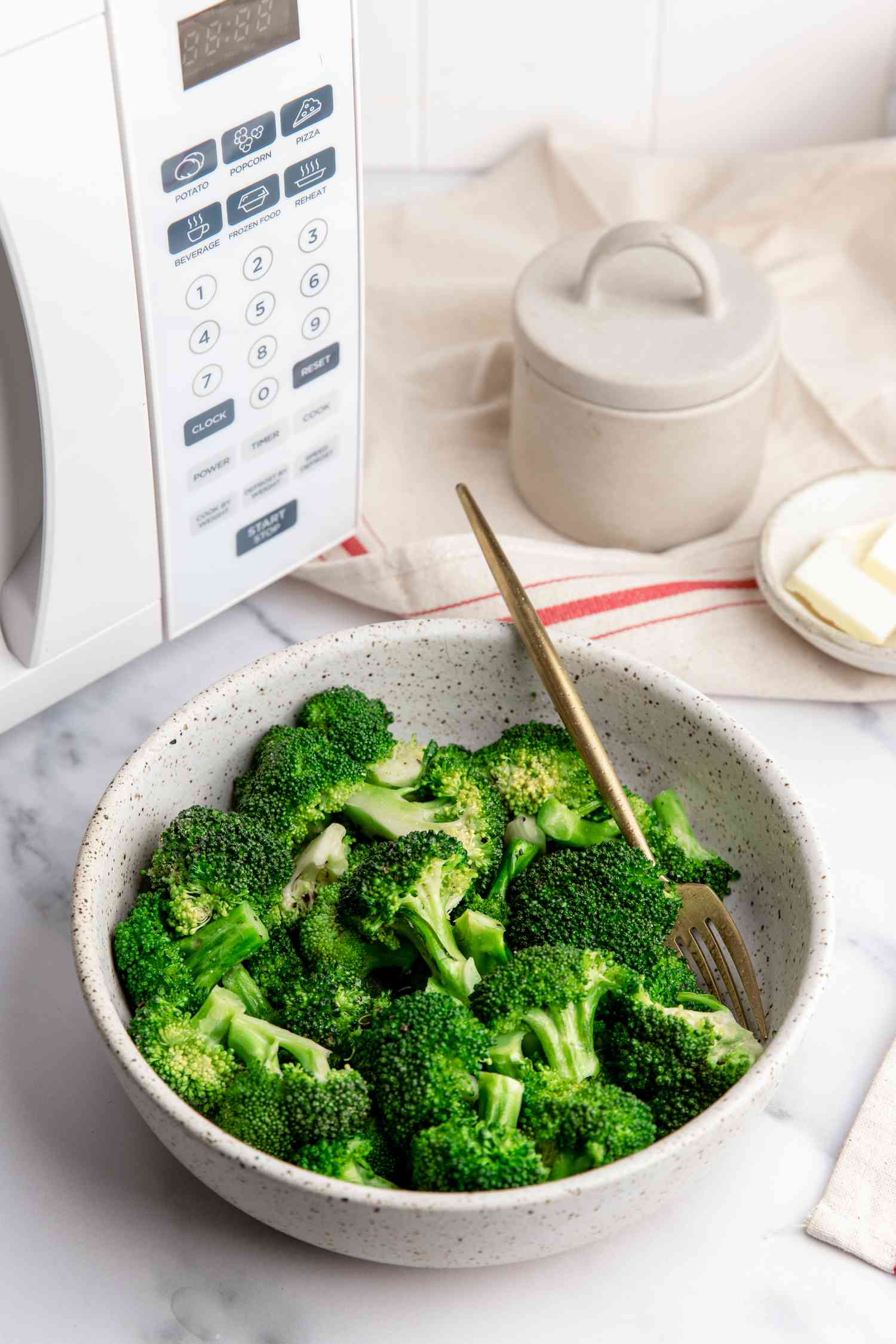 Bowl of Microwave Steamed Broccoli with a Fork, and in the Background, a Counter Setting with a Microwave, a Container of Salt, a Small Saucer with Slices of Butter, and a Kitchen Towel