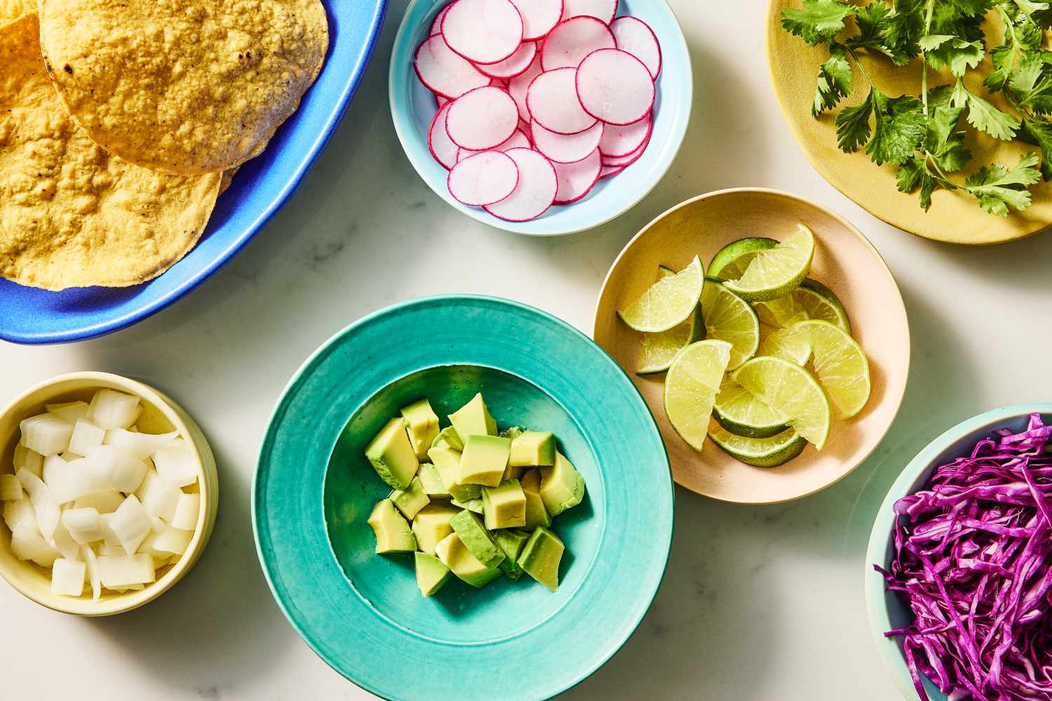 Ingredients for a dish including tortillas radishes lime wedges cilantro diced onions avocado chunks and shredded purple cabbage on a table