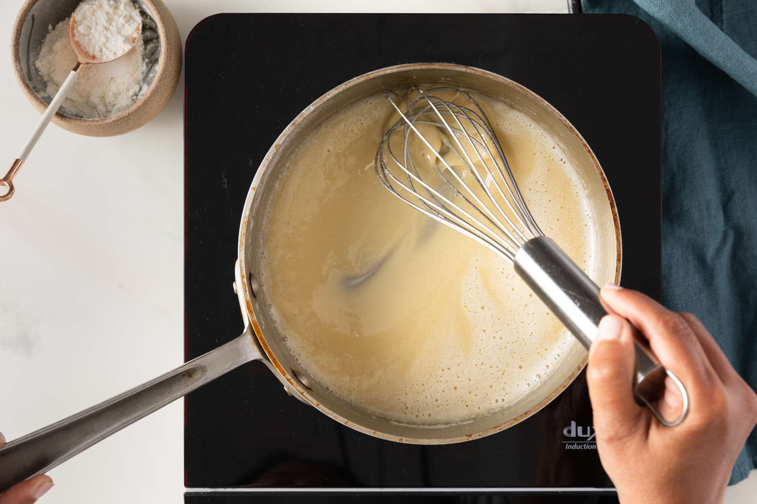 Flour well whisked with the melted butter in a saucepan, almost at a white roux (pot on a portable stovetop next to a bowl of flour and a blue kitchen towel) for how to make roux