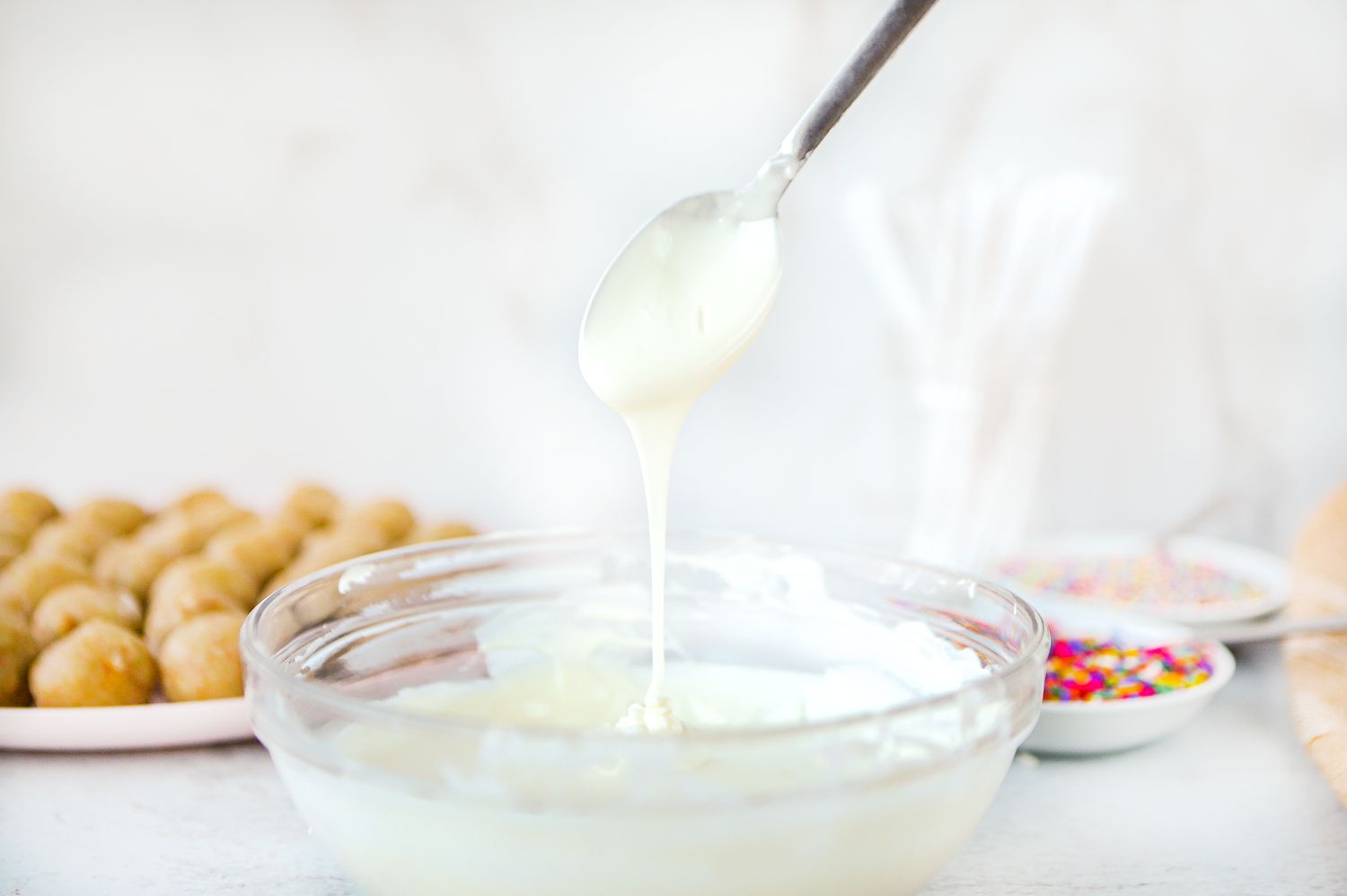 White Chocolate in a Bowl Scooped with a Spoon with Cake Pops and Sprinkles in Background