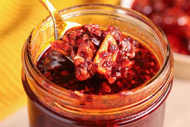Spoonful of Homemade Chili Crispy Raised from Mason Jar With More Crisp, and in the Background, a Saucer With More Chili Crisp and a Kitchen Towel