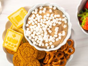 A bowl of hot cocoa dip topped with marshmallows surrounded by cookies pretzels and crackers on a plate