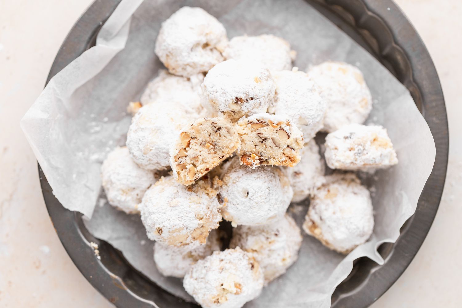 Overhead view of walnut snowball cookies piled on a parchment lined decorative baking pan with the top one cut in half to show the interior.