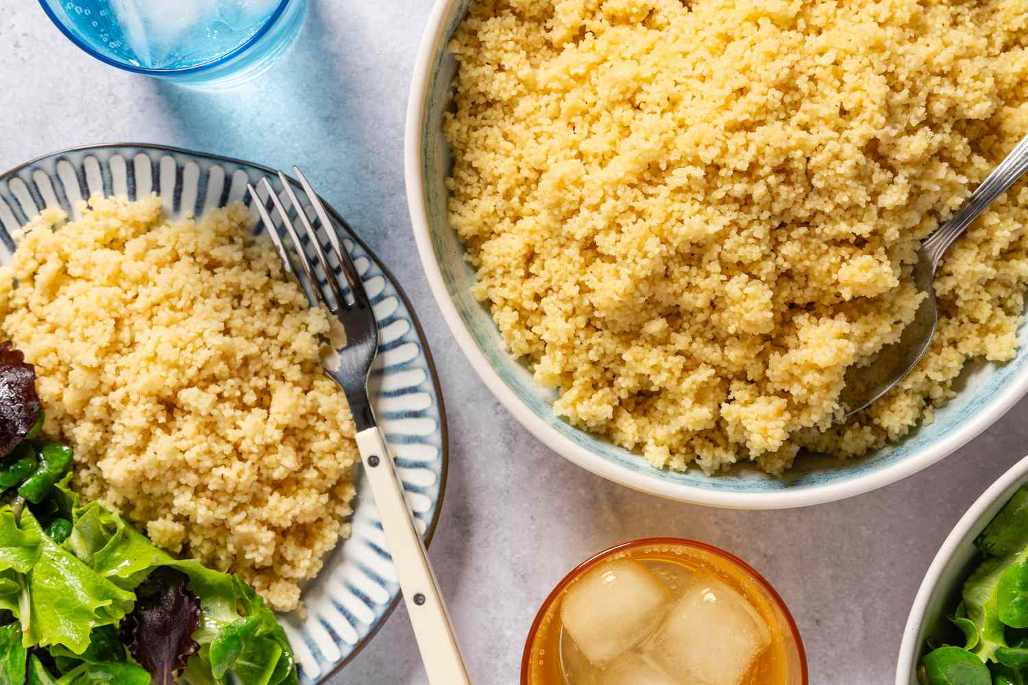 Overhead view of a large bowl of Cheesy Couscous with a serving spoon next to a serving plate with fork and lettuce
